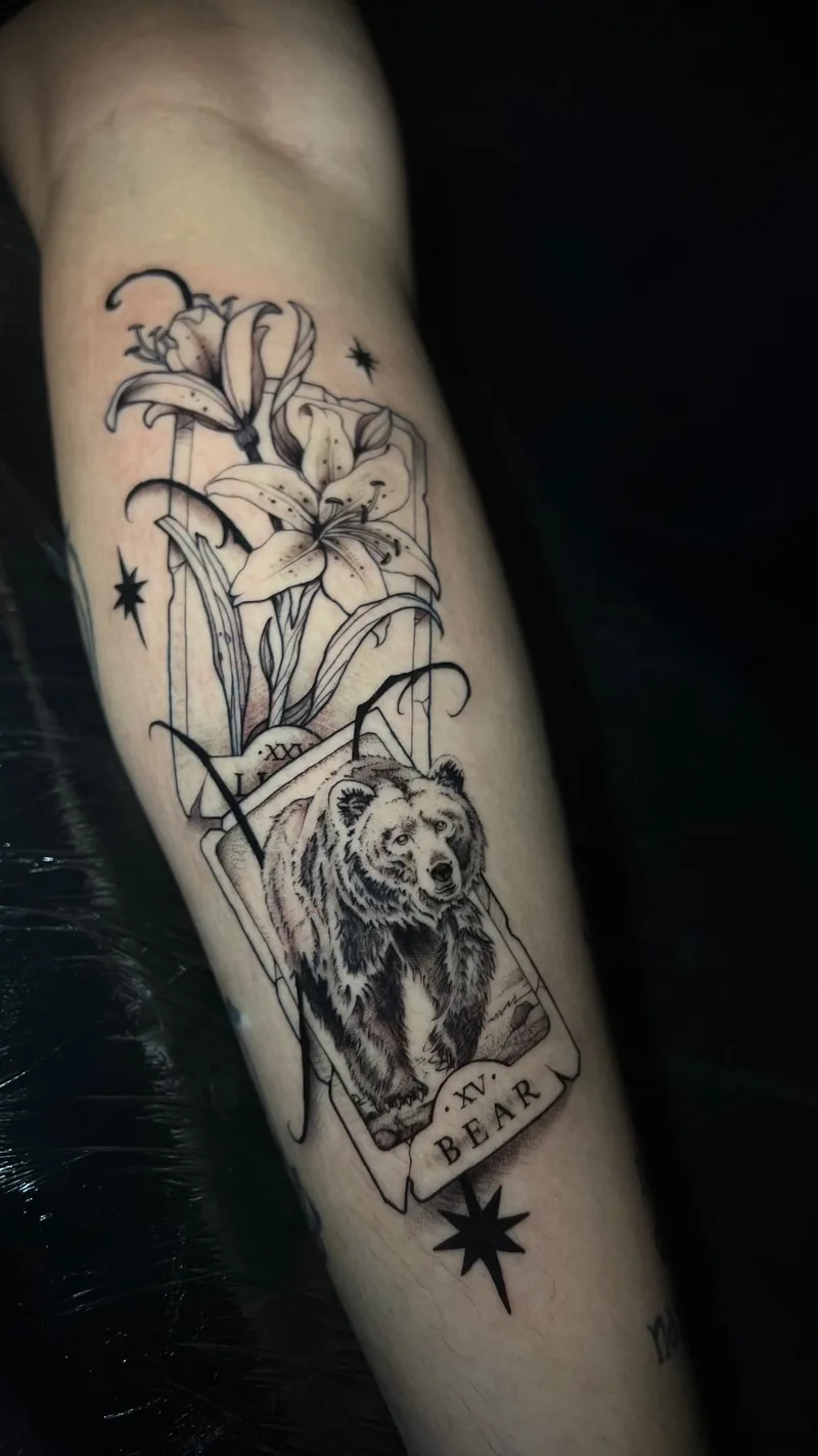 Tatuagem em tinta preta de urso em uma carta, com lírios, faixas e estrelas ao redor